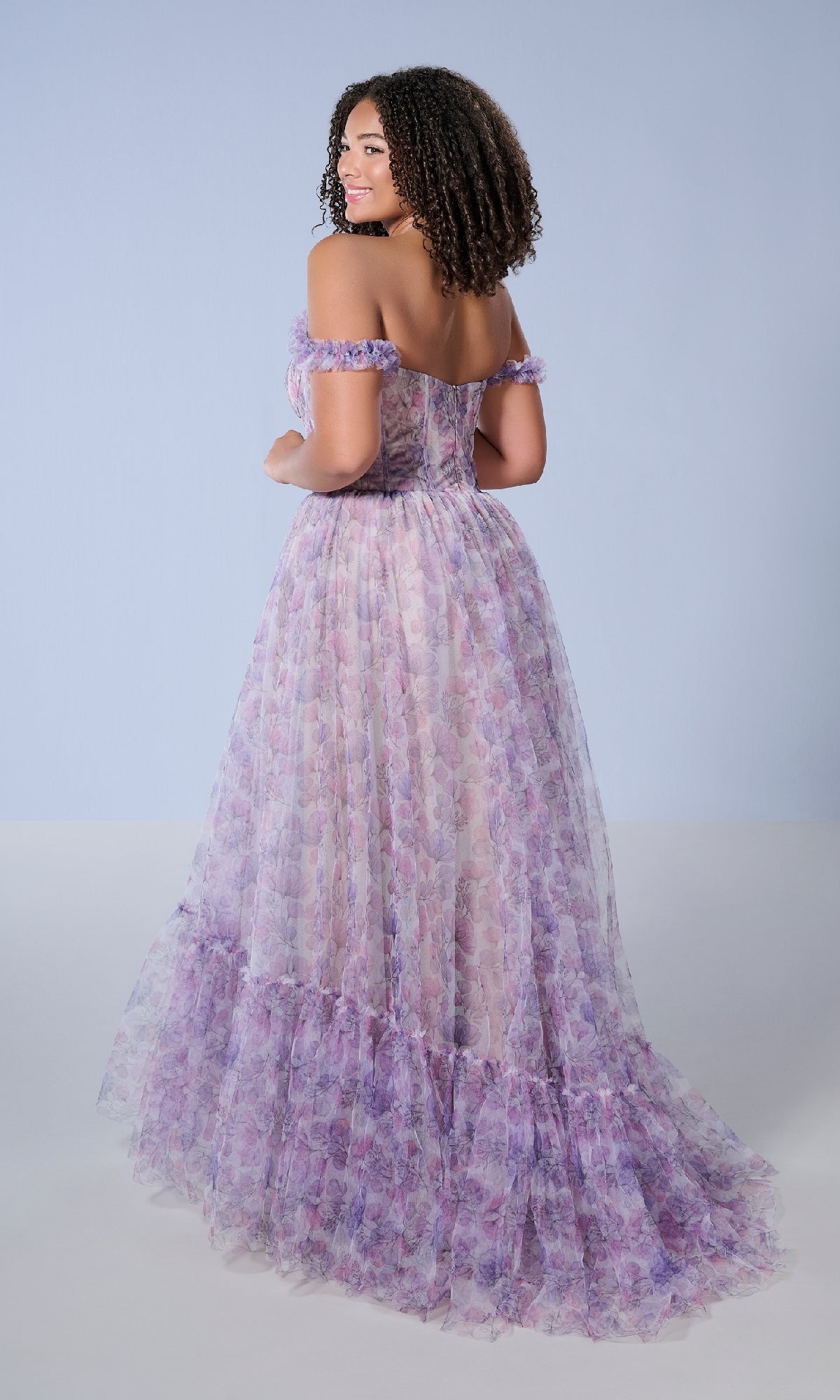 A woman wears the Tiffany 16162 Long Plus-Size Prom Dress—an off-shoulder, floor-length lavender floral gown with a corset bodice—posing with her back turned and looking over her shoulder against a light blue background.